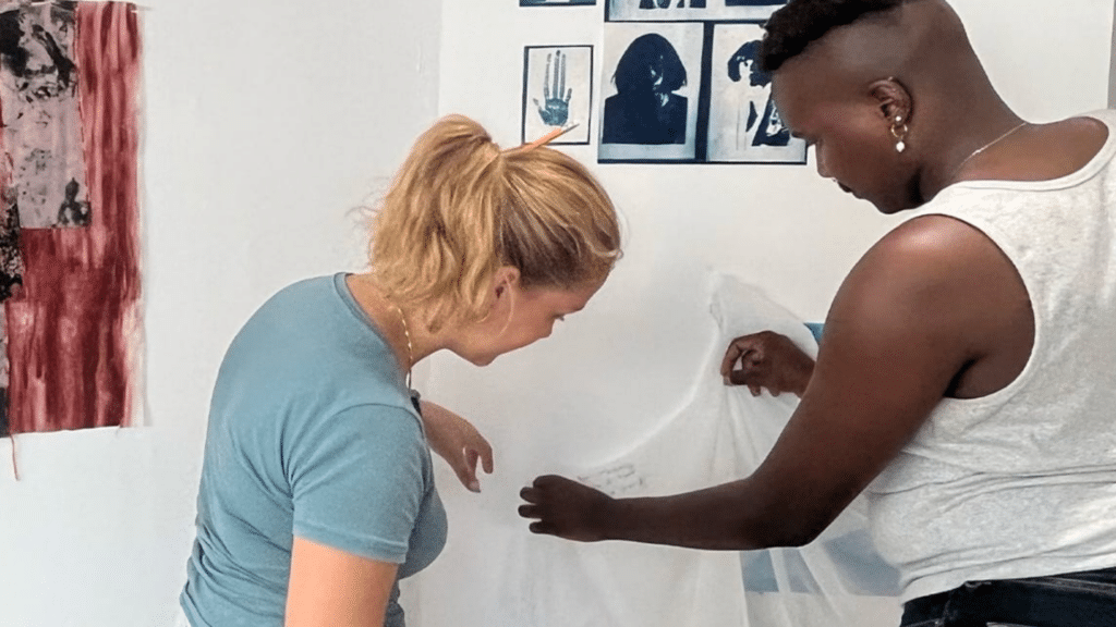 Two people in a bright room work with white fabric; one seated, one standing, smoothing the material.