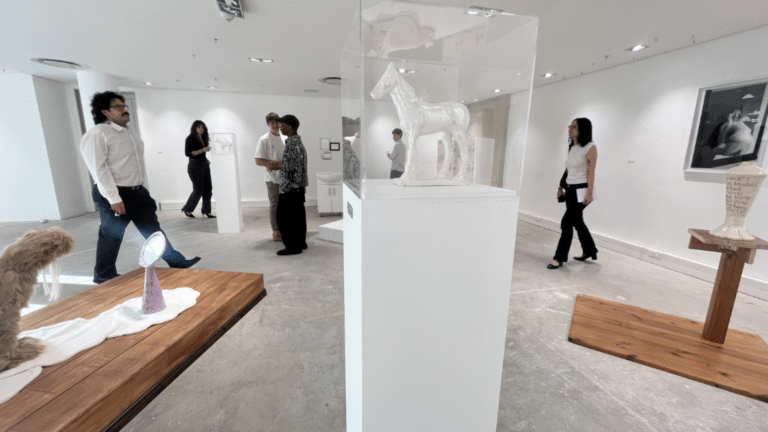 Visitors and students walking through a professional white-cube gallery space featuring sculptures and framed photography.