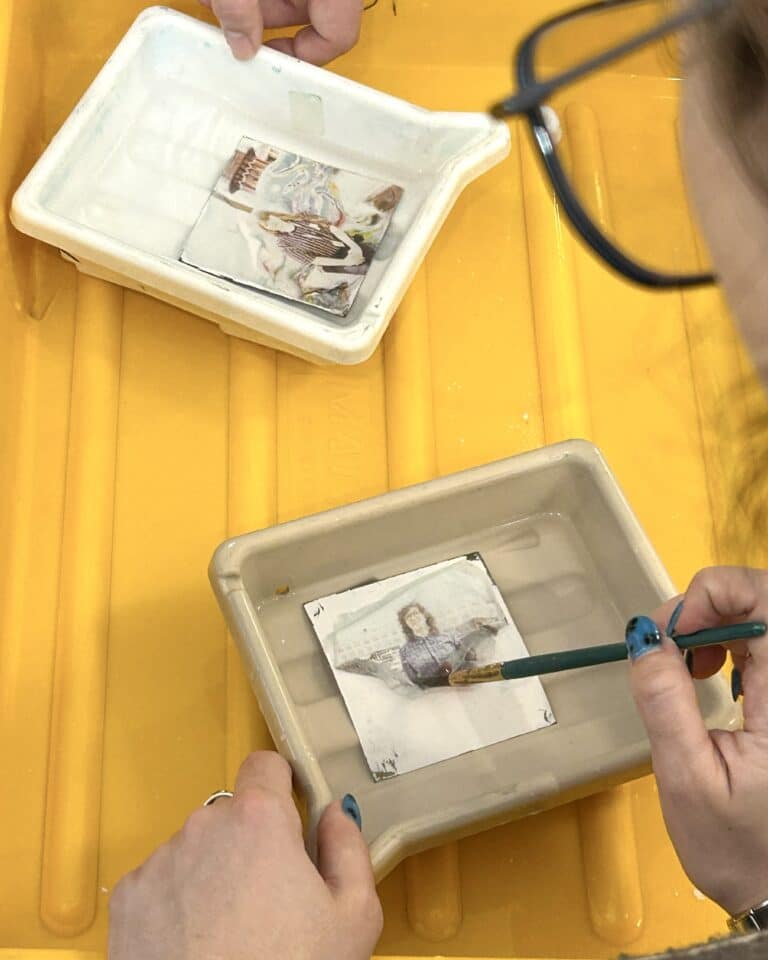 Hands mosaic-assembling small tiles over a white tray on a bright yellow surface; a person holds a pencil with blue paint marks nearby, indicating an art project.