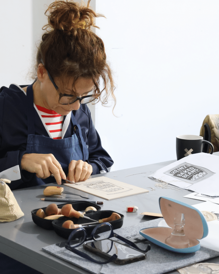 Woman wearing glasses and a blue jacket sits at a table, cutting paper with crafting tools scattered around.