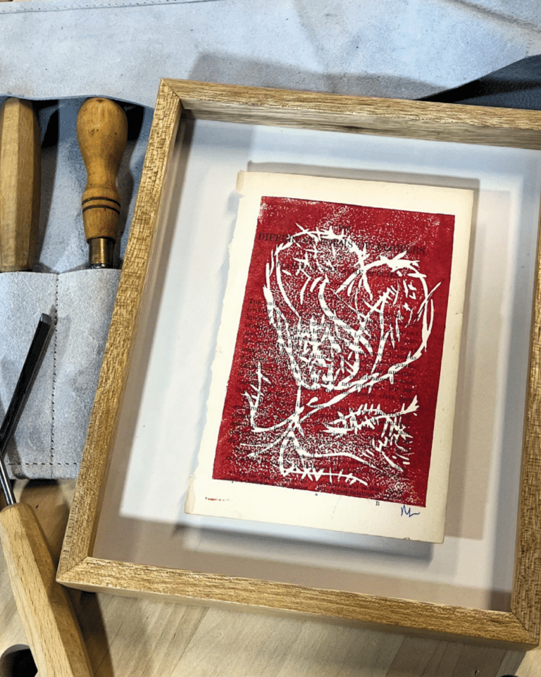 Framed red graphic print with white tree design, set in a light wooden frame on a white mat, with wooden tool handles nearby.