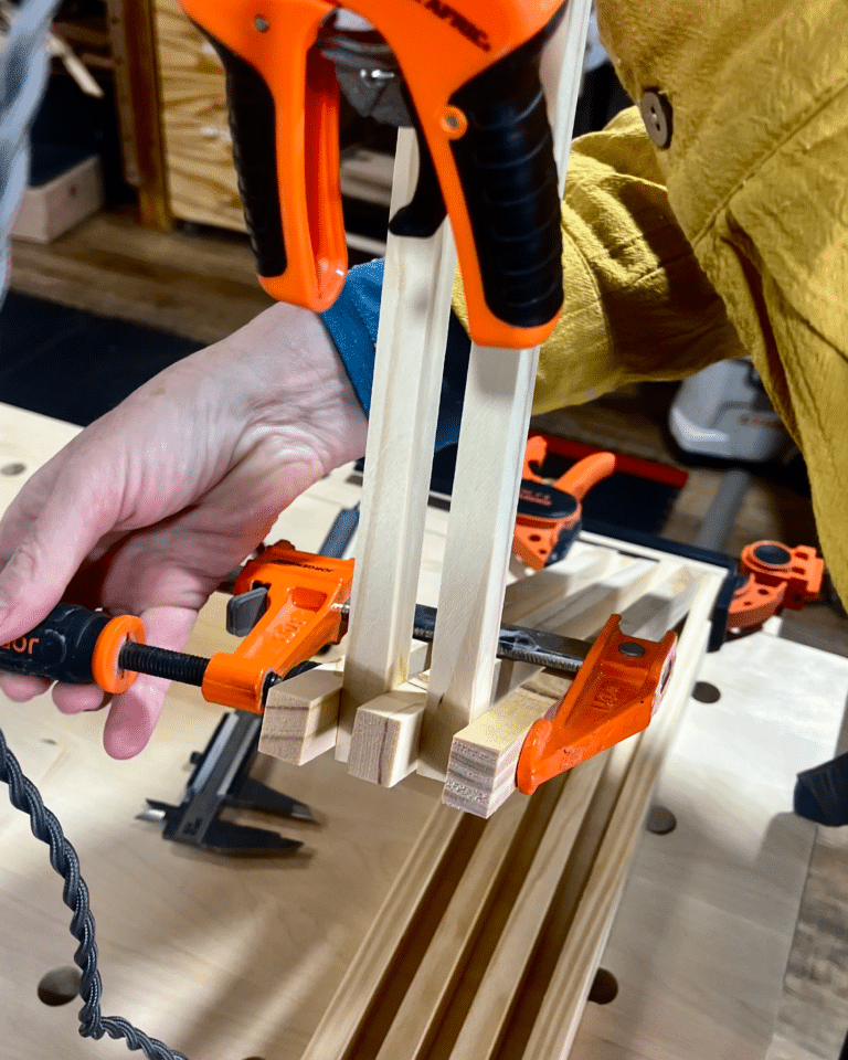 Close-up of orange bar clamps securing thin wooden slats in a woodworking project, with a hand holding the pieces in place.