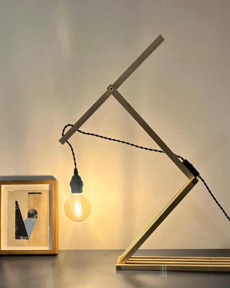 Geometric metal frame sculpture with an exposed edison bulb hanging from a twisted cord, against a neutral wall with a framed artwork below.