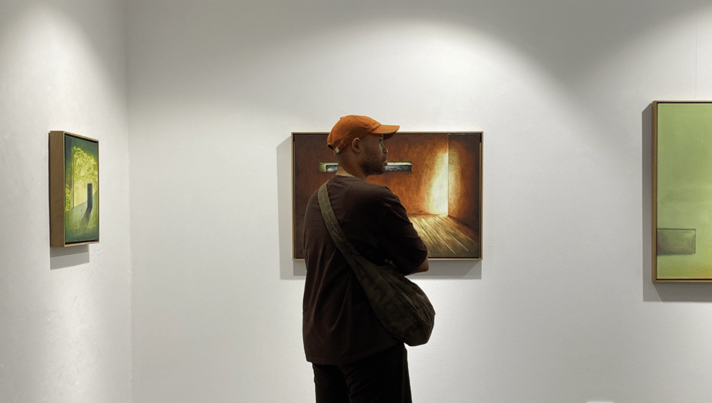 Man in an orange cap and brown jacket stands in a gallery, looking at a large brown-toned painting on the wall.