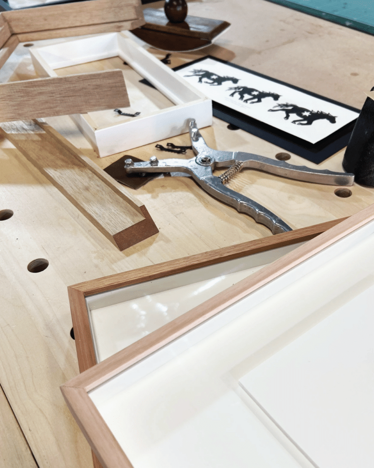 Woodworking bench with wooden picture frames, a metal clamp, and a sheet of black animal silhouettes nearby on the work surface.