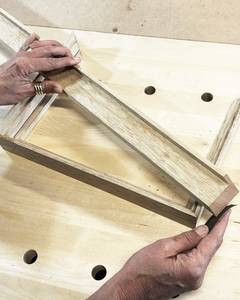 Hands with rings fit a wood corner joint on a light plywood frame atop a workbench.